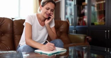 Smiling woman talking on phone and writing notes in notebook. Note taking and telecommuting