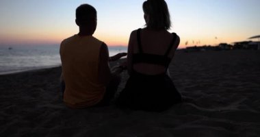 Couple in love watching sunset on beach together and traveling on summer vacation. Silhouette of people from behind sitting enjoying sunset sea view