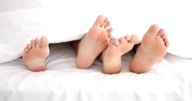Bare feet of loving couple under white blanket on bed. Family intimate life