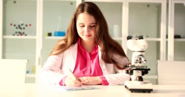 Beautiful female scientist uses microscope and writes in notebook in laboratory. Scientific research goals and objectives