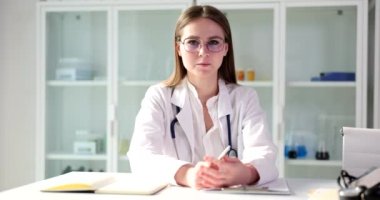 Doctor waving smiling looking at camera and sitting at table in clinic. Nurse therapist video call with patient or record video blog with medical advice