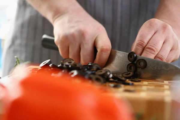 stock image Cook cuts black ripe olives with knife on board. Useful and dangerous properties of olives in dish and salad