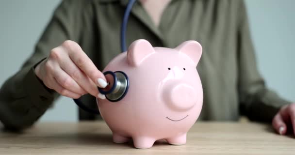 手握听诊器听粉色储蓄罐与硬币近距离4K电影慢动作 健康保险费用概念 — 图库视频影像