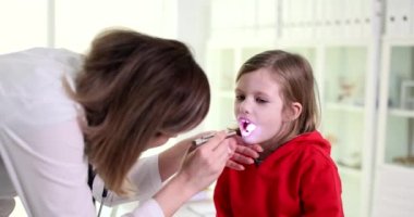 Kadın otolaringolog doktor, acı sendromlu hasta çocuğun boğazına ışık tutuyor. Çocukta büyümüş bademcik ve akut bademcik iltihabı.