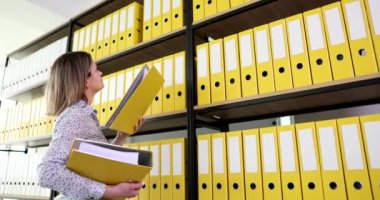 Muhasebeci kadın arşiv belgelerinin olduğu sarı klasörü rafa koyar. Belgeler ve finansal raporlama örgütüyle çalışmak