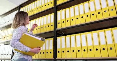 Kadın sekreter, ofis 4K filminde ağır çekimde üst raftan sarı klasörü çıkarıyor. İş konseptinde bürokrasi