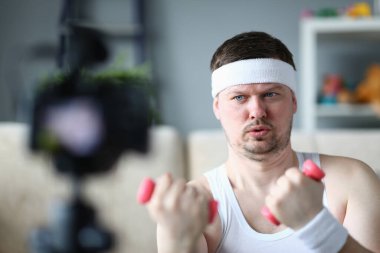 Atletik adam kamerada dambıllarla fiziksel egzersiz yapıyor. Uzak çevrimiçi fitness ve erkek blog yazarı
