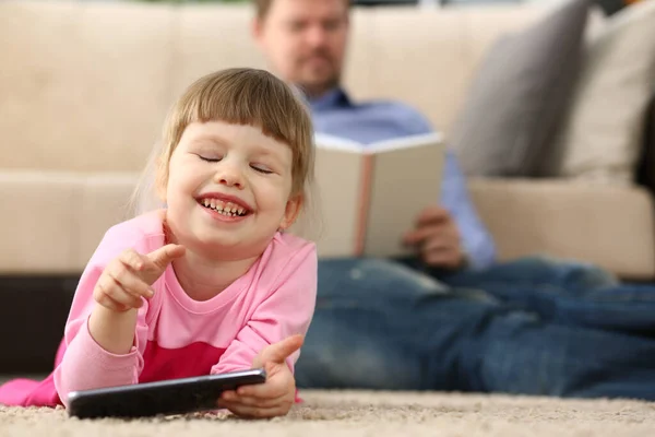 Little smiling baby girl with smartphone lies on floor in background dad reads a book. Children interests in phone and reading books