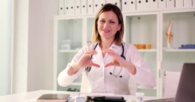 Mutlu kadın masada oturan bir doktor kalbin işaretini gösterir, yakın plan. Pozitif duygular, tıp