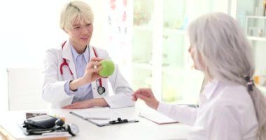 Doktor endokrinolog, klinikteki yaşlı kadın hastaya ağır çekimde yeşil elma veriyor. Yaşlılık kavramında rasyonel beslenme