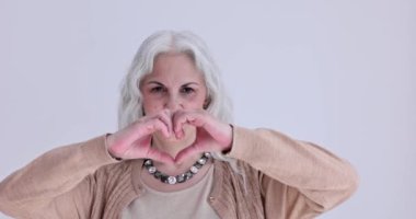 Olgun bir kadın, ışık stüdyosunda gülümseyen avuçlarıyla kalp gösterir. Güzel gri saçlı bayan beyaz arka planda yavaş çekimde flört ediyor. Aşk ilişkisi