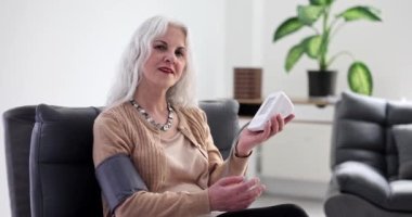 Mutlu olgun kadın evde yavaş çekimde kan basıncını dijital tonometreyle ölçer. Sağlık durumu teşhisi için ekipman. Hasta kişisel bakım.