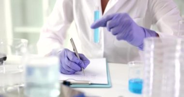 Bilimadamı laboratuvarda incelenen kimyasal reaktörün rengini ve durumunu tanımlıyor. Lastik eldivenli kadın elinde mavi bir sıvıyla ağır çekimde oturuyor.