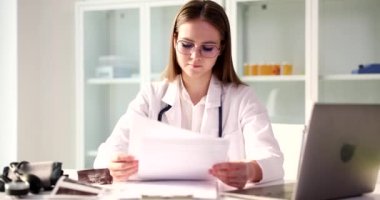 Odaklanmış kadın doktor, açık dizüstü bilgisayarın yanındaki masada oturan hastanın test sonuçlarına bakıyor. Kadın, klinikte ağır çekimde tıbbi evrakları inceliyor.