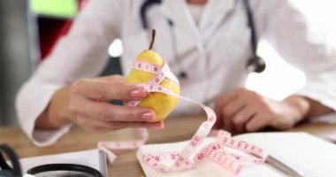Doktorun elinde bantla sarılı taze armut var. Klinikteki masada oturuyor. Tıp üniformalı kadın olgun meyve gösteriyor. Sağlıklı diyet kavramı