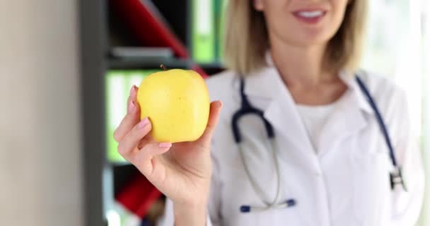 正の女性医師はクリニックで笑顔手に新鮮なリンゴを保持しています 医療制服の陽気なブロンドの女性は健康的な食事をお勧めします — ストック動画
