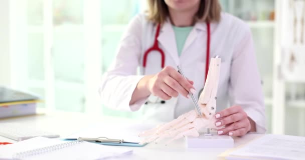 女性はテーブルに座って骨格手首の人工モデルの骨を指しています 聴診器を装着した医師が人体のスローモーションの構造を調べる — ストック動画