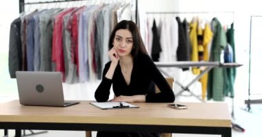 Pozitif genç bir kadın açık dizüstü bilgisayarın yanında oturuyor ve kameraya bakıyor. İş yerindeki yorgun esmer yönetici yavaş çekimde kıyafetlere karşı.