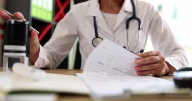 Kadın doktor hastane ofisindeki masada duran evrakları zımbalıyor. Steteskoplu bir kadın klinikte ağır çekimde hasta geçmişi ile çalışıyor.