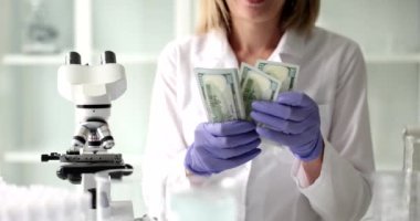 Bilim adamı elektronik mikroskobun yanında duran dolar banknotlarını saydı. Laboratuvar önlüklü kadın bilimsel araştırmalardan sonra maaşını kontrol ediyor.