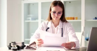 Odaklanmış doktor, dizüstü bilgisayarına bakan hasta materyallerini inceliyor. Gözlüklü genç kadın tıp tarihi okuyor. Masada ağır çekimde oturuyor.