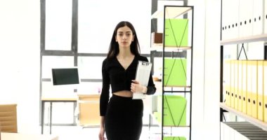 Modern ofiste elinde pano olan şık esmer kadın kameraya bakıyor. Resmi kıyafetli ciddi bir bayan yönetici ağır çekimde çalışmaya hazır.