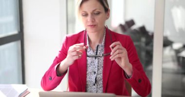 Odaklanmış kadın işe başlamadan önce gözlük takar ve saçlarını eliyle düzeltir. Pembe ceketli müdür açık dizüstü bilgisayarın yanında ağır çekimde oturuyor.