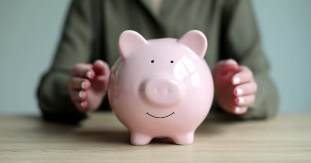 女人把双手放在粉红储蓄罐的侧面 硬币拥抱着木桌 爱钱的容器用于储蓄和积存慢动作 — 图库视频影像