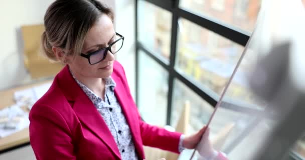 オフィスの大きな窓の近くに立つホワイトボードには 眼鏡をかけた集中型の女性がメモを書きます スマートマネージャーは仕事のスローモーションで戦略を開発 — ストック動画