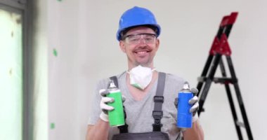 Mutlu ustabaşı yeşil ve mavi şişelerden beyaz boya püskürtür. Koruyucu miğferli ve gözlüklü neşeli adam yavaş çekimde gülümsüyor.
