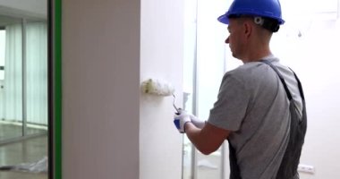 Mavi kasklı, odaklanmış ustabaşı beyaz duvarları silindirle boyuyor. İş kıyafetli bir adam daireyi yavaş çekimde yenilemek için özel bir araç kullanıyor.