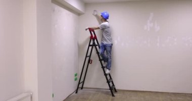 Foreman, tadilat sırasında duvarın sıvanmış halini kontrol etmek için merdivenin üzerinde duruyor. İş kıyafetli adam ağır çekimde gülümseyen jestler gösteriyor.