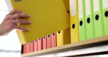 Sekreter sarı klasörde malzeme rafına geri koyar. Arşiv dosyaları yavaş çekimde olan çok renkli halka klasörleri