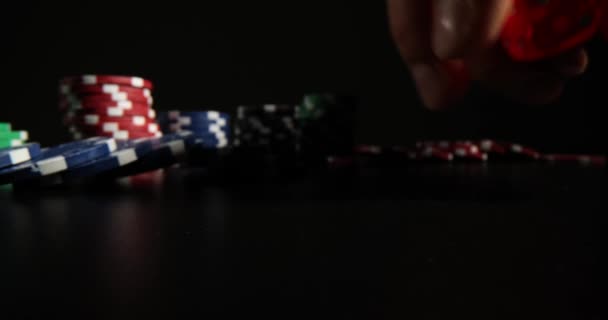 男はカラフルなポーカーチップの近くの暗いテーブルの表面に赤いサイコロをスローします 黒の背景にプロのギャンブルゲームをプレイするための機器 — ストック動画