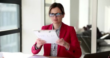 Takım elbiseli ve gözlüklü, meşgul, üzgün bir kadın elinde kağıt levhalarla ofiste oturuyor. Ofis işi ve iş yükü kavramı