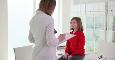 Dosyası olan çocuk doktoru çocuk kızı klinikte muayene ediyor. Çocuk doktoru hasta kızla konuşuyor.