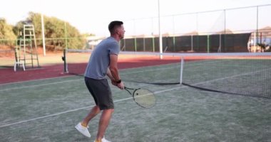 Tenisçi sert kortta raketle vuruyor. Spor kıyafetli genç adam güneşli bir günde tenis oynuyor.