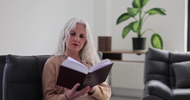 高齢の白髪の女性はソファでリラックスして本を読んでいます 高齢者生活における読書の役割 — ストック動画