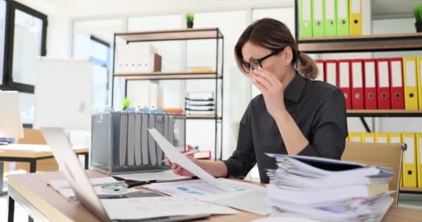 疲れた女社員がノートパソコンで机に座っている紙を見て眼鏡を外します オフィスワーカーが職場で業務データを確認するのを心配 — ストック動画