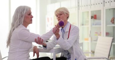Bakıcı doktor, kıdemli hastanın ağırlığıyla klinikte dirsek bükmesine yardımcı oluyor. Yetenekli ortopedist, beyaz saçlı kadınların eklemlerini yavaş çekimde kontrol ediyor.
