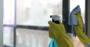 Lastik eldivenli biri deterjan sıkar ve pencere camlarını yavaş çekimde siler. Kadın evde temizlik malzemeleriyle yüzeyi yıkıyor.