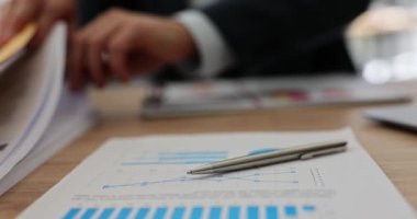 Kalem, ofis masasında belge arayan müdüre karşı açılan soruşturma kağıdının üzerinde. Yavaş çekimde istatistik ve grafiklerle muhasebe malzemeleri
