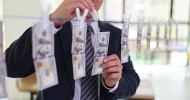 İşadamı, çamaşır ipini iğneyle sabitlemek için dolar banknotları asıyor. Klasik takım elbiseli başarılı bir adam ofiste ağır çekimde para aklıyor.