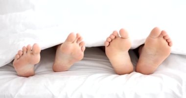 Çıplak ayaklı adam, sabahları yatakta yatan kadına dokunur. Çıplak ayaklı evli çift yatak odasında beyaz battaniyeden sarkıyor.