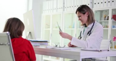 Gülümseyen kadın çocuk doktoru, iş yerindeki küçük hastanın test sonuçlarına bakıyor. Kadın ve kız klinikteki masada ağır çekimde oturuyor.
