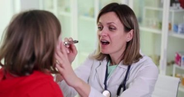 Odaklanmış doktor, parlayan el feneriyle hasta kızın boğazını kontrol etmek için ağzını daha geniş açmasını istiyor. Üniformalı profesyonel doktor ağır çekimde sağlık kontrolü yapıyor.