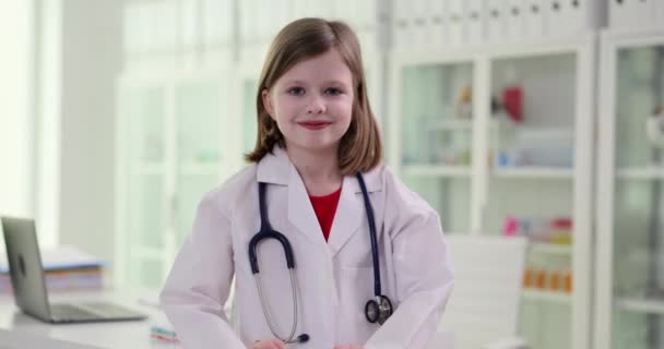 白いコートを着た中学生は医師の役割を真似ており 心臓の兆候を示しています ポジティブな女の子とともにStethoscopeスタンドで現代オフィス遅い動き — ストック動画