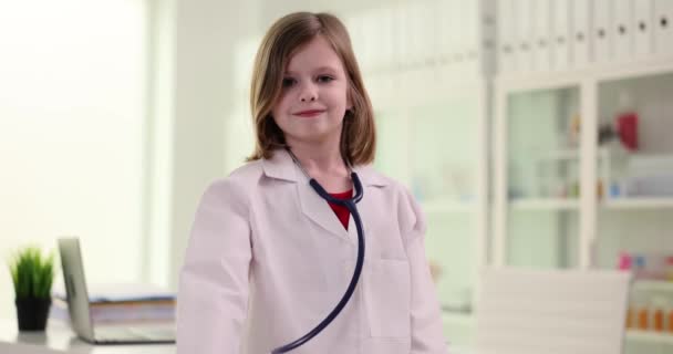 白衣を着た笑顔の中学生は医師の役割を模倣し 聴診器を示しています ポジティブな女の子スタンドでライトドクターオフィススローモーション — ストック動画