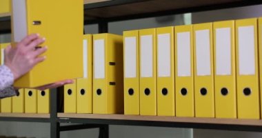 Kadın çalışan, muhasebe belgeleriyle birlikte sarı yüzük klasörünü ofis arşivine geri koyuyor. Rafta malzemeler olan organize dizin yapısı