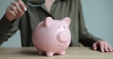 Kadın eli, pembe domuz kumbarasının yanına büyüteç koyuyor. Kadın, gülümseyen domuz kumbarasının içine bakmaya çalışır ve birikmiş parayı ağır çekimde görür.